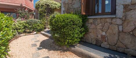 A stone building with a window and a door, surrounded by gravel and greenery.