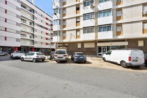 Un immeuble à plusieurs étages, avec plusieurs voitures garées et un fourgon blanc devant.