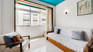 Una habitación con sofá, una silla, ventana con cortinas, una impresión botánica enmarco y techo con detalles azules.