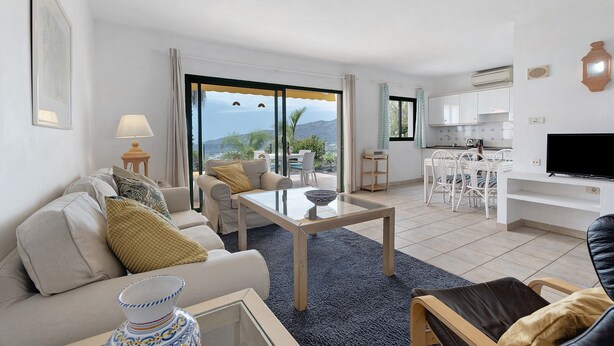 A living room with a sofa, coffee table, and a TV. There is a balcony with a view of the sea. - Holiday Home 'La Villa La Palma- 2 Dormitorios' with Sea View, Wi-Fi and Air Conditioning (Dos Pinos)