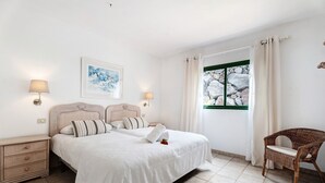 Un dormitorio ordenado con una cama, dos mesitas de noche, una silla, una ventana con cortinas y un cuadro enmarcado en la pared.