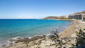 Una spiaggia con un litorale roccioso, un mare blu cristallino e un albergo a più piani sullo sfondo.