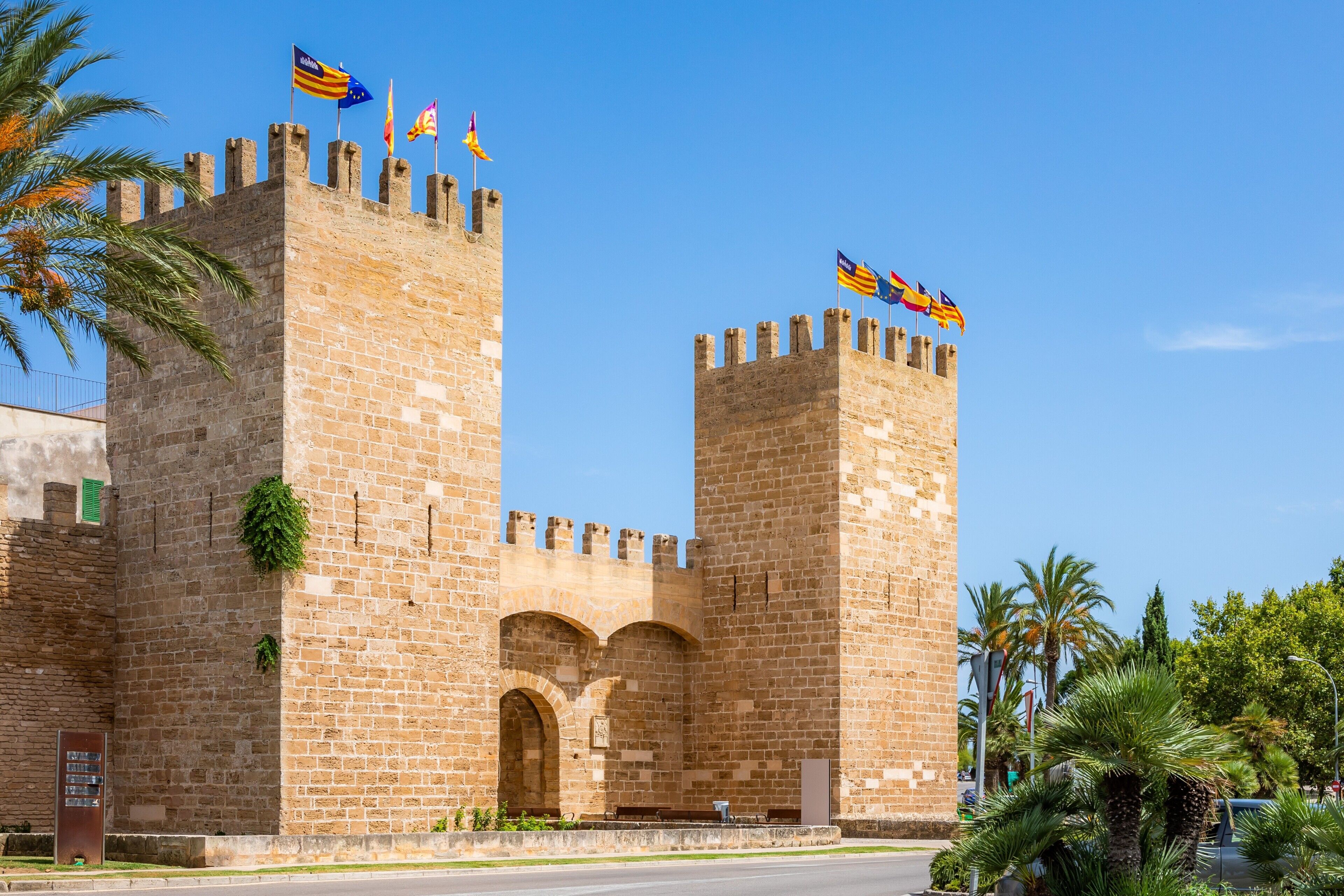A historic stone castle with two prominent towers, a central arched entrance, and flags on top.