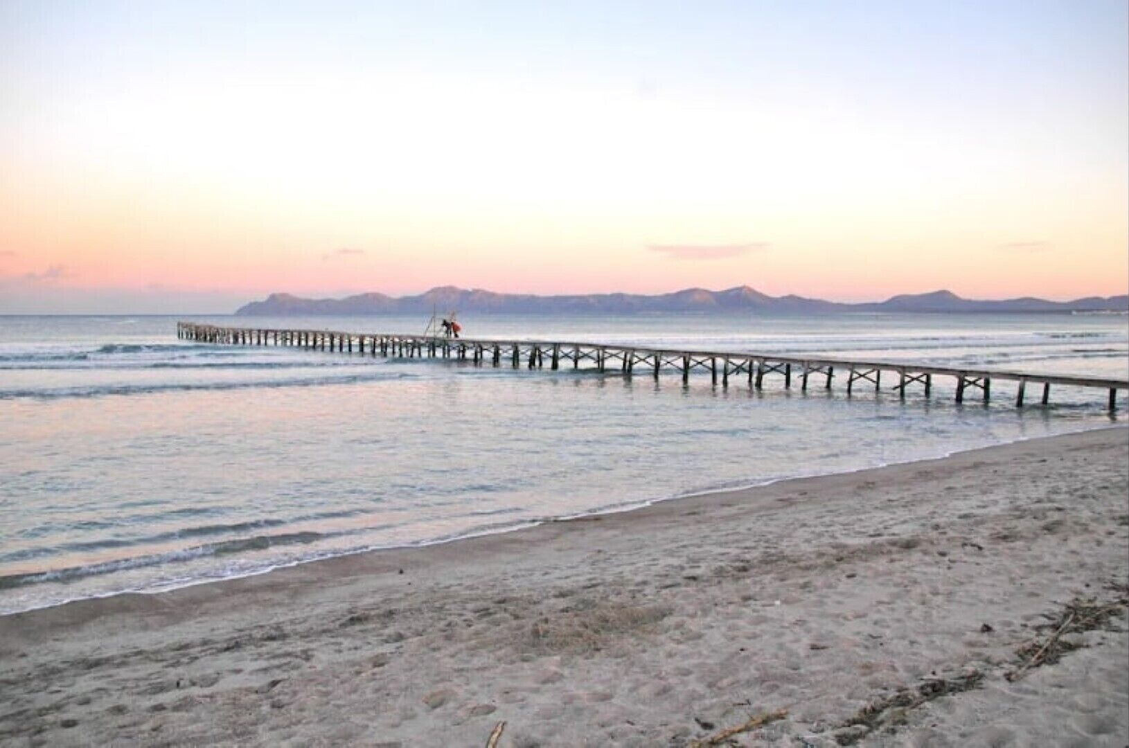 A beach with a wooden pier extending into calm waters, a clear sky, and distant mountains.