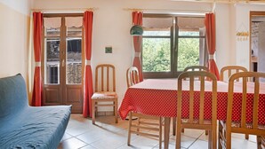 A room with a wooden ceiling, a dining table with a red tablecloth, wooden chairs, a blue sofa, and a window with red curtains.