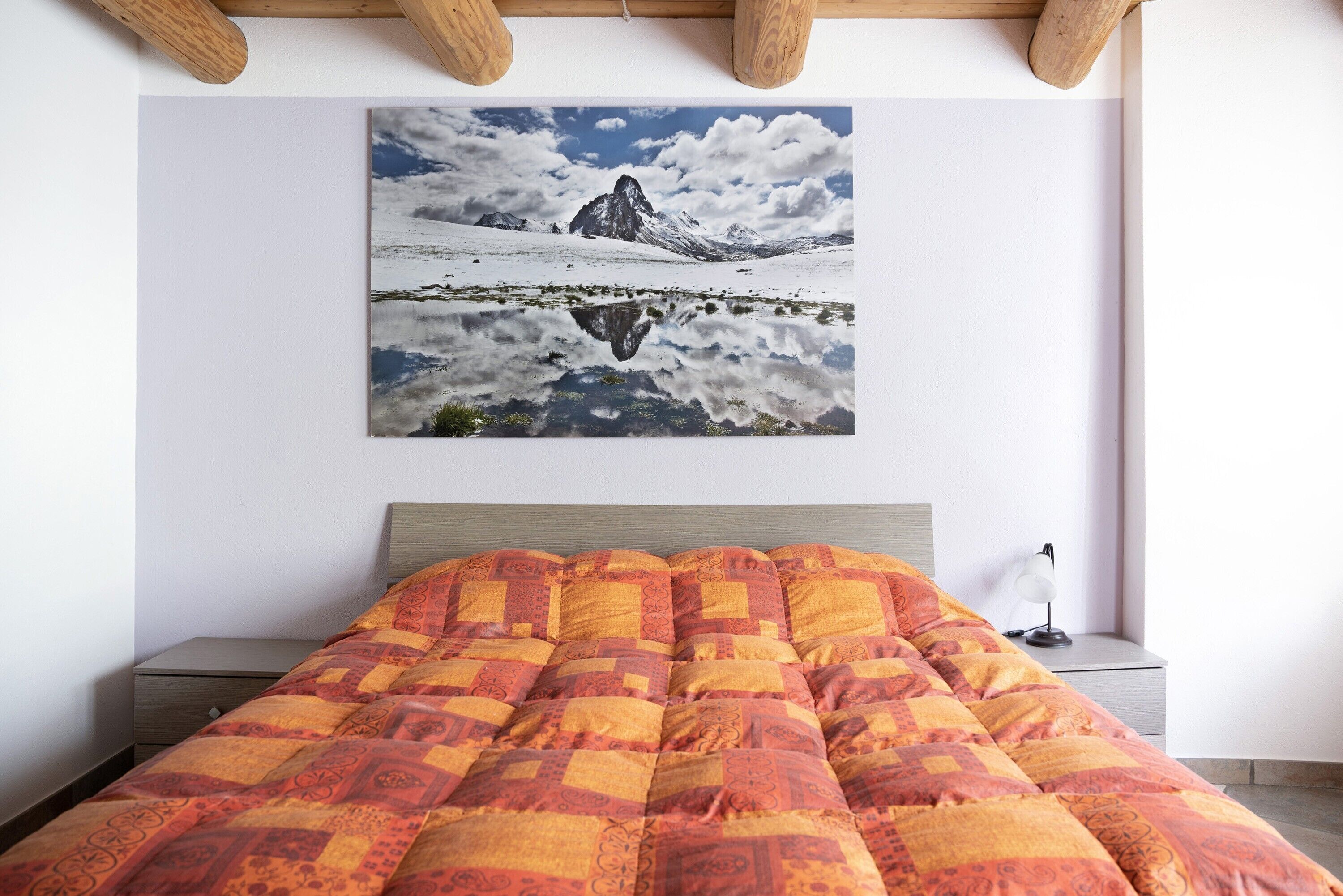 Un lit recouvert d’une couverture à motifs, une lampe de chevet et, au mur, un tableau représentant un paysage de montagne.