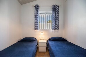 Une petite chambre avec deux lits, une fenêtre ornée de rideaux à motifs, une table de chevet avec une lampe, et une vue sur les palmiers à l’extérieur.