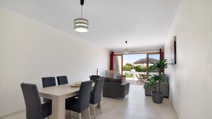 Un área de comedor con una mesa y sillas de madera, un sofá, una planta en maceta y vistas al exterior a través de una puerta de vidrio.