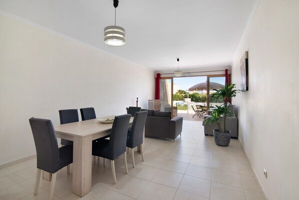 Uma área de refeições com uma mesa e cadeiras de madeira, um sofá, uma planta em vaso e vista para uma área externa através de uma porta de vidro.