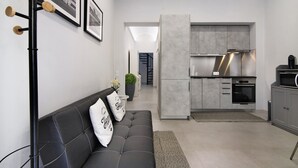 Una sala moderna con un sofá negro, una mesa de centro y un mueble de pared. Se observa una zona de cocina con armarios grises y electrodomésticos de acero inoxidable.