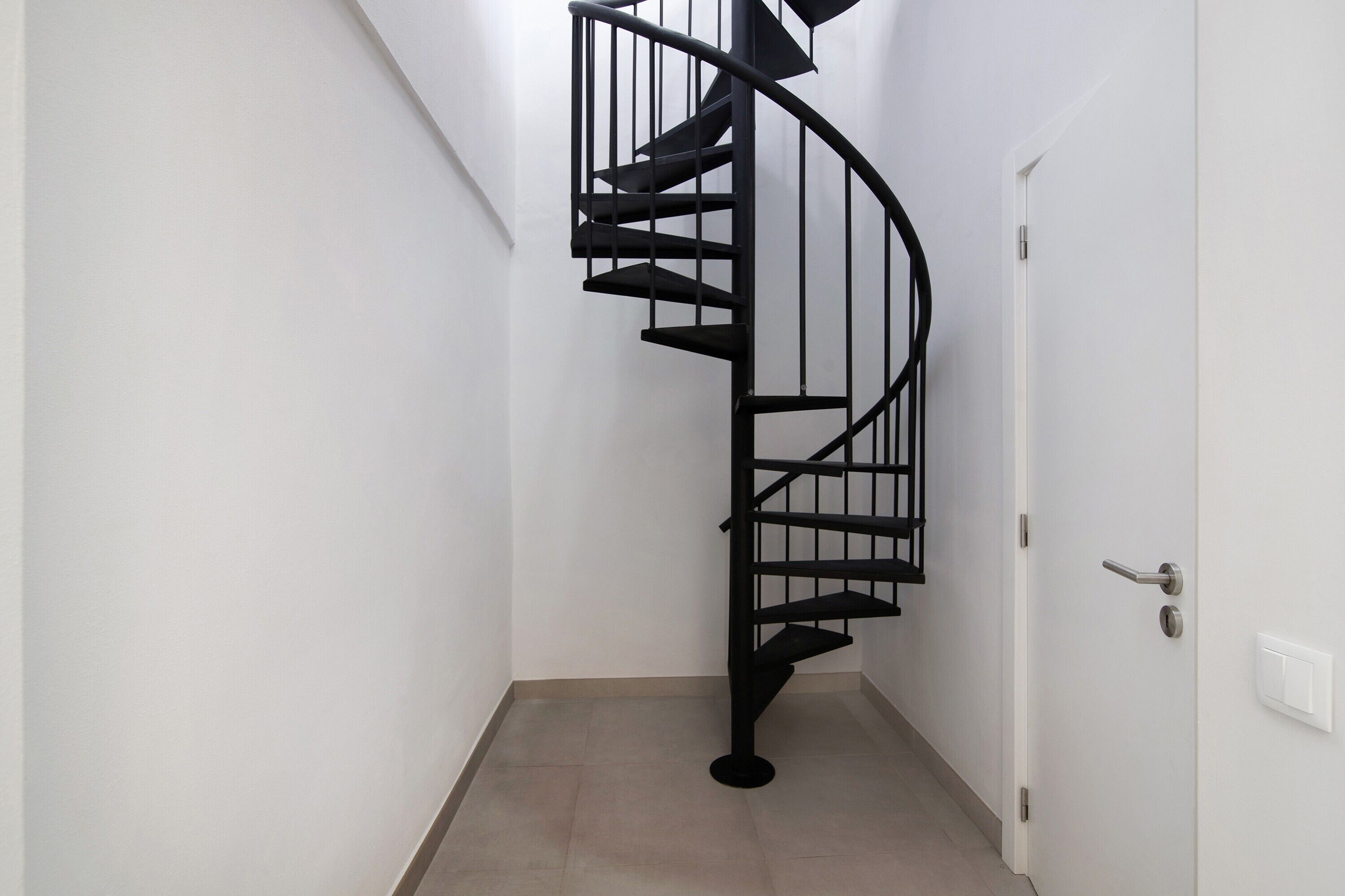Una escalera en espiral en un pasillo con paredes blancas y una puerta con picaporte plateado.