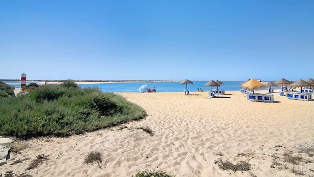 Una playa con sombrillas de paja, sillas de descanso y un faro al fondo.