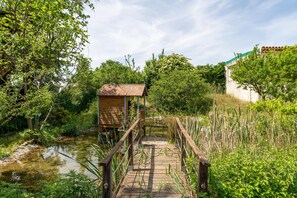 A wooden bridge leading to a rustic wooden structure with a tiled roof, surrounded by lush greenery and a calm water body.