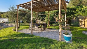 Una pérgola de madera con una mesa y sillas debajo, rodeada de un jardín cuidado.