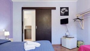 Habitación de hotel con una cama, un escritorio con computadora, un televisor y una puerta que da a otra habitación.