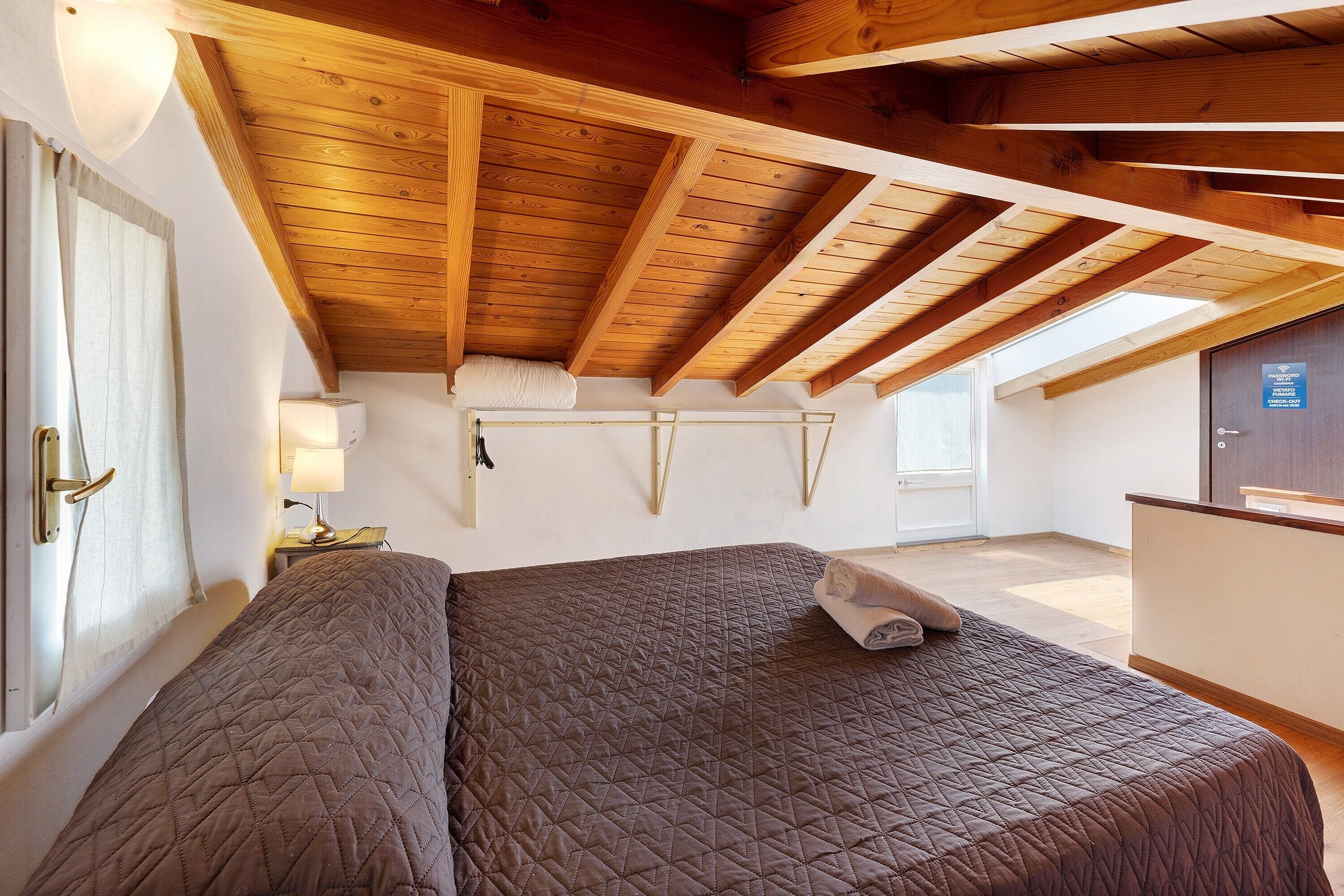 Een slaapkamer met een houten plafond, een bed met een gewatteerde spreidek en een nachtlampje, en een raam met gordijnen.