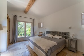 A bedroom with a bed, a nightstand, a window with curtains, and a wooden ceiling beam.