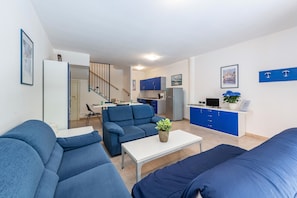 A modern living room with blue sofas, a white coffee table, and a kitchen in the background.