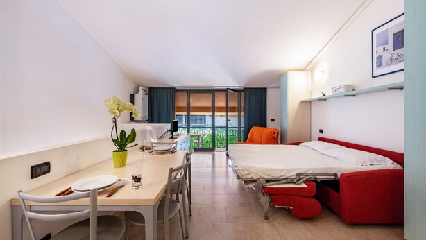 Habitació d'hotel senzilla amb zona de dinar, llit amb calaixos d'emmagatzematge, planta en test i balcó amb vistes.