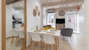 Un comedor con una mesa blanca, sillas blancas y un sofá gris. Se observa una cocina al fondo con armarios blancos y una encimera negra.