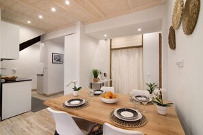 Uma sala de jantar com uma mesa de madeira, cadeiras brancas, uma cesta de frutas e um vaso de planta.
