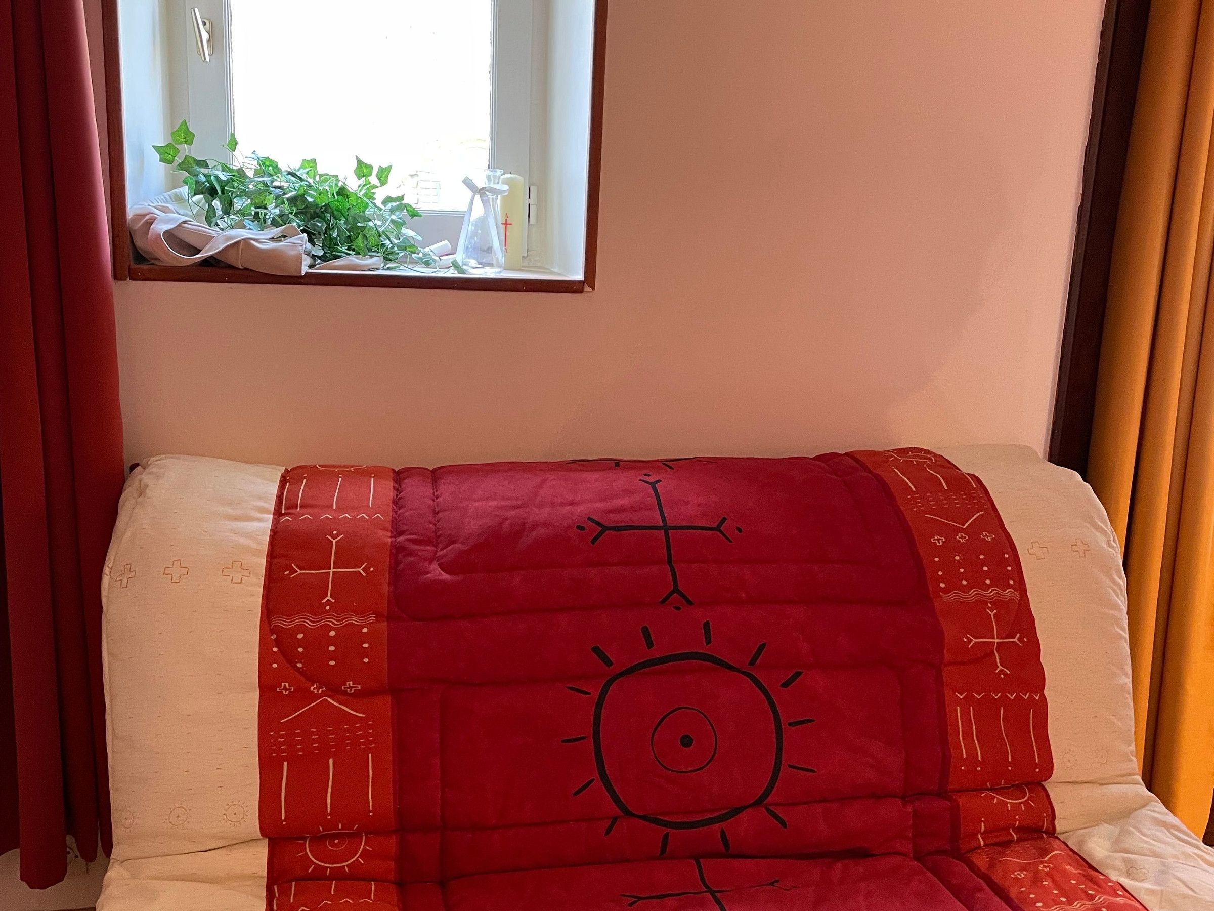Un lit recouvert d’une couverture rouge ornée d’un motif représentant un soleil, une fenêtre avec des rideaux et une plante posée sur le rebord de la fenêtre.