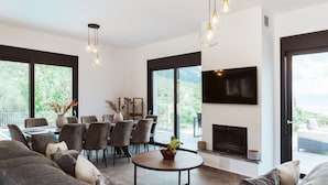 Een moderne woonkamer met een open haard, een eethoek en grote ramen met uitzicht op groen.