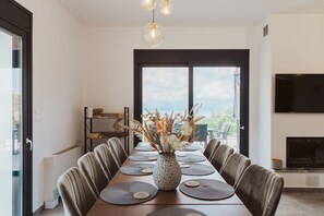 Un coin repas avec une longue table dressée pour six personnes, un vase rempli de fleurs séchées et une vue sur l’extérieur grâce à de grandes fenêtres.