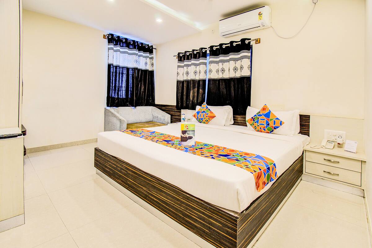 Una habitación de hotel con una cama grande, una mesita de noche, un equipo de aire acondicionado y una ventana con cortinas.