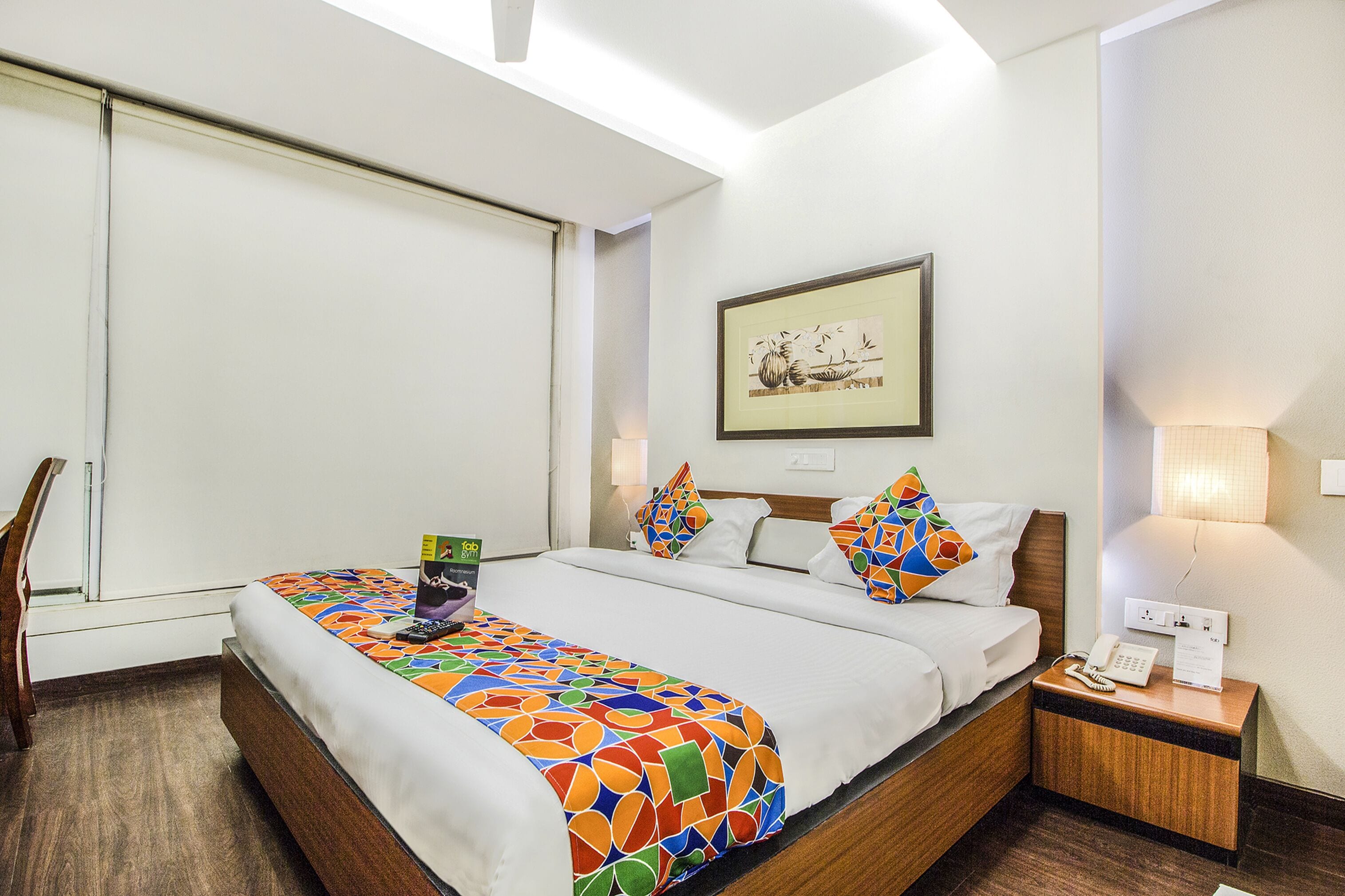 A hotel room with a bed, a nightstand, a lamp, a framed picture on the wall, and a window with blinds.