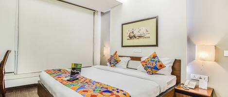 A hotel room with a bed, a nightstand, a lamp, a framed picture on the wall, and a window with blinds.