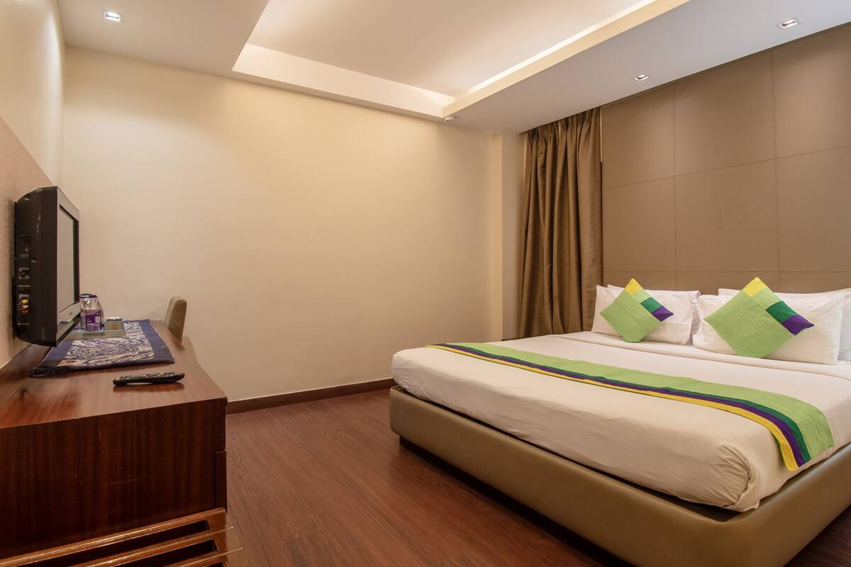 A hotel room with a bed, a desk with a flat-screen TV, and a control panel.