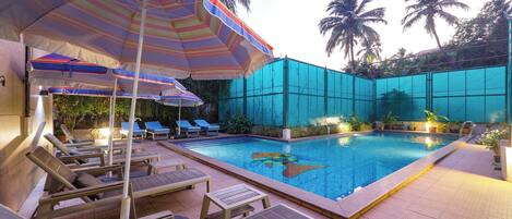 A swimming pool with lounge chairs and umbrellas, surrounded by a fence and palm trees.