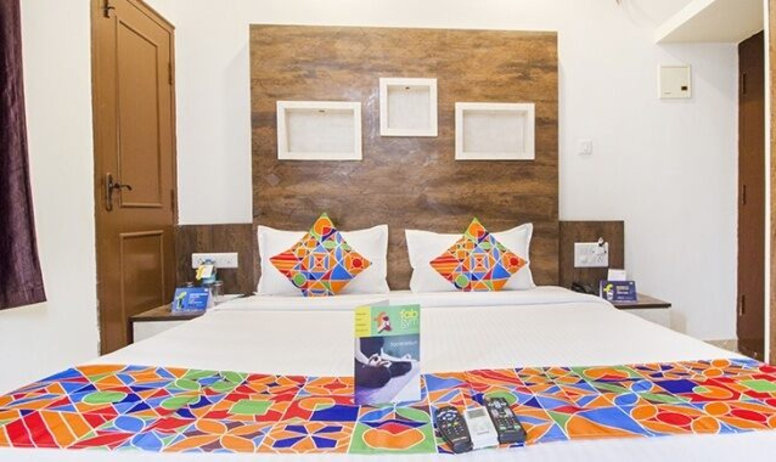 A hotel room with a wooden headboard, two pillows with colorful geometric patterns, a bed with a colorful mosaic bedspread, and a remote control on the bed.