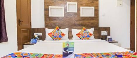 A hotel room with a wooden headboard, two pillows with colorful geometric patterns, a bed with a colorful mosaic bedspread, and a remote control on the bed.