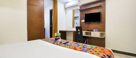 Una habitación de hotel con cama, escritorio, silla, televisión y un estante pequeño.