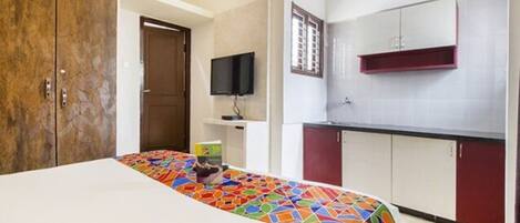 A bedroom with a bed, a TV, and a kitchenette with cabinets and a window.