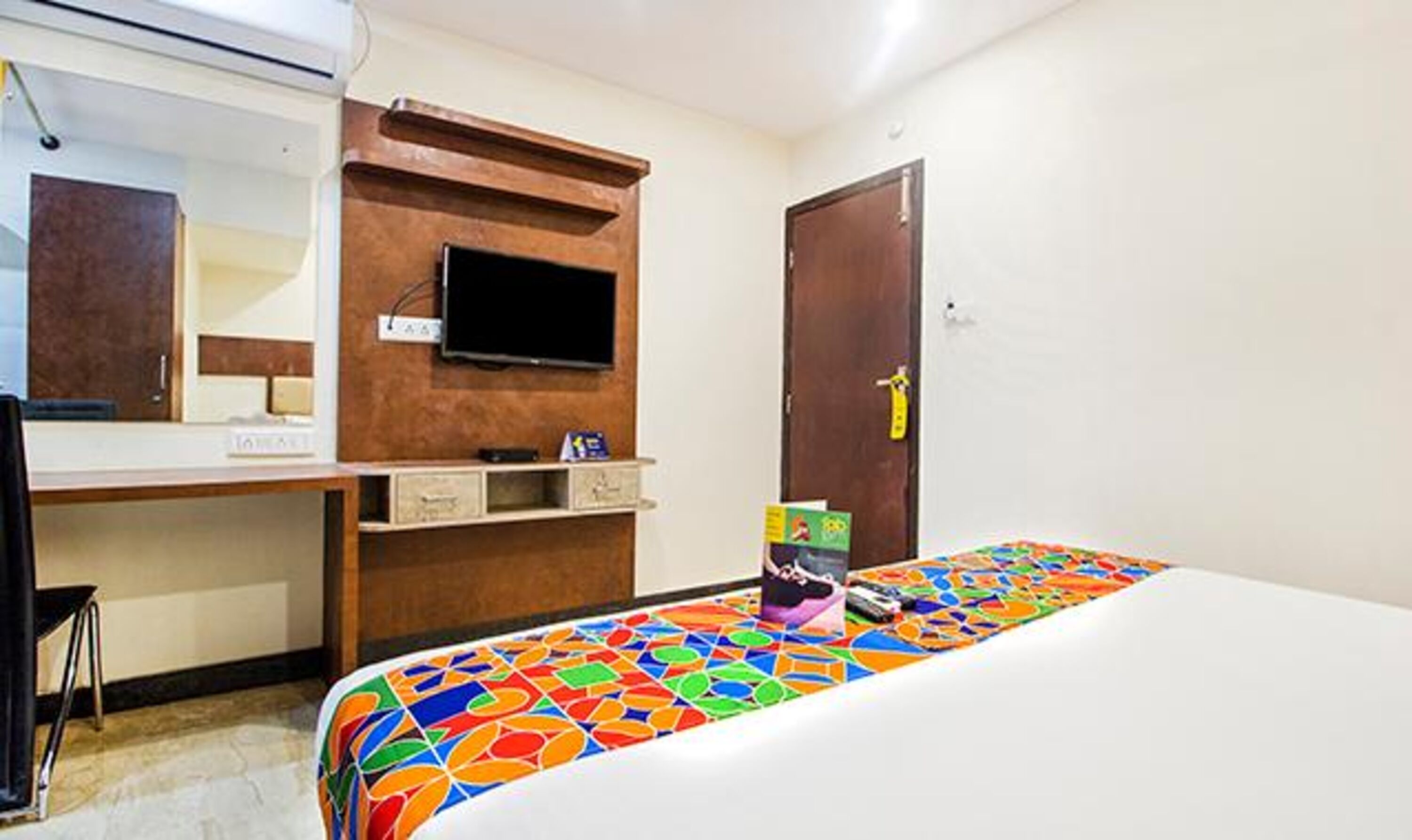 Una habitación de hotel con una cama, un televisor montado en la pared, un aparador de madera y una puerta con picaporte amarillo.