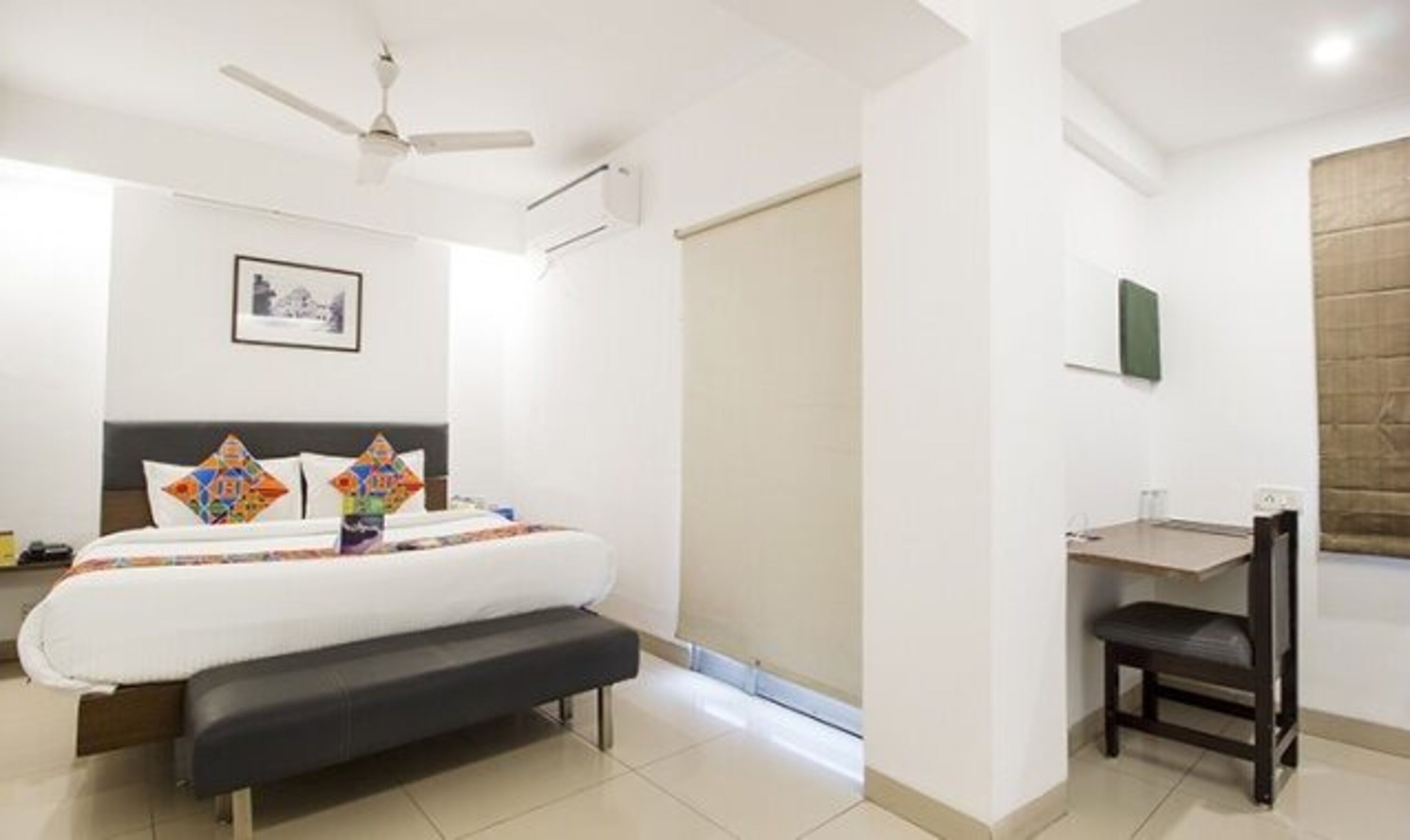 A hotel room with a bed, a desk, a chair, a ceiling fan, and a framed picture on the wall.