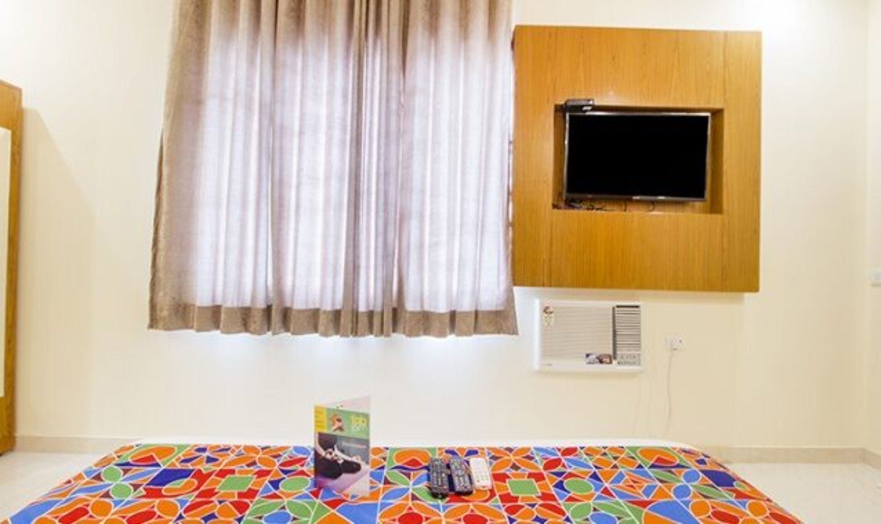 Een kamer met een bed, een raam met gordijnen, een wandtelevisie en een airconditioner.