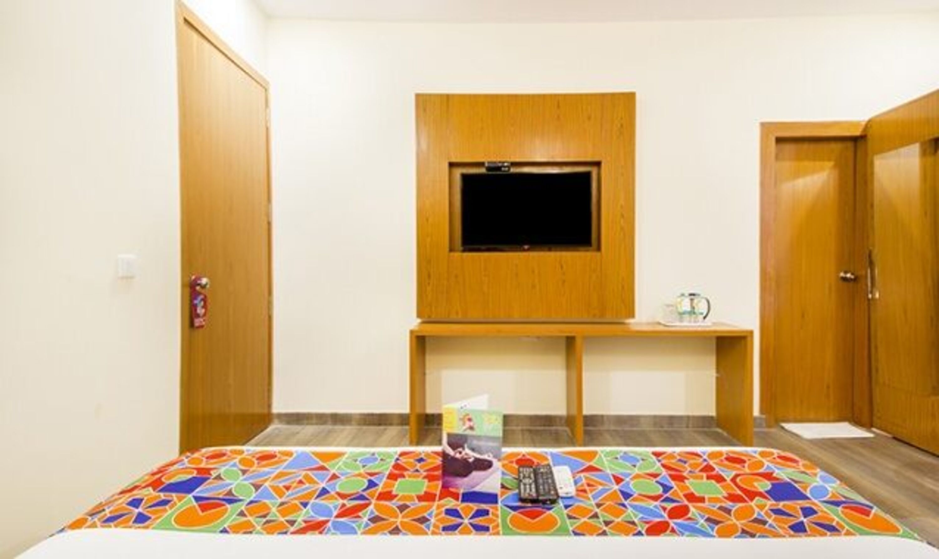 A room with a bed, a TV mounted on a wooden stand, a small table with a kettle, and a colorful rug.