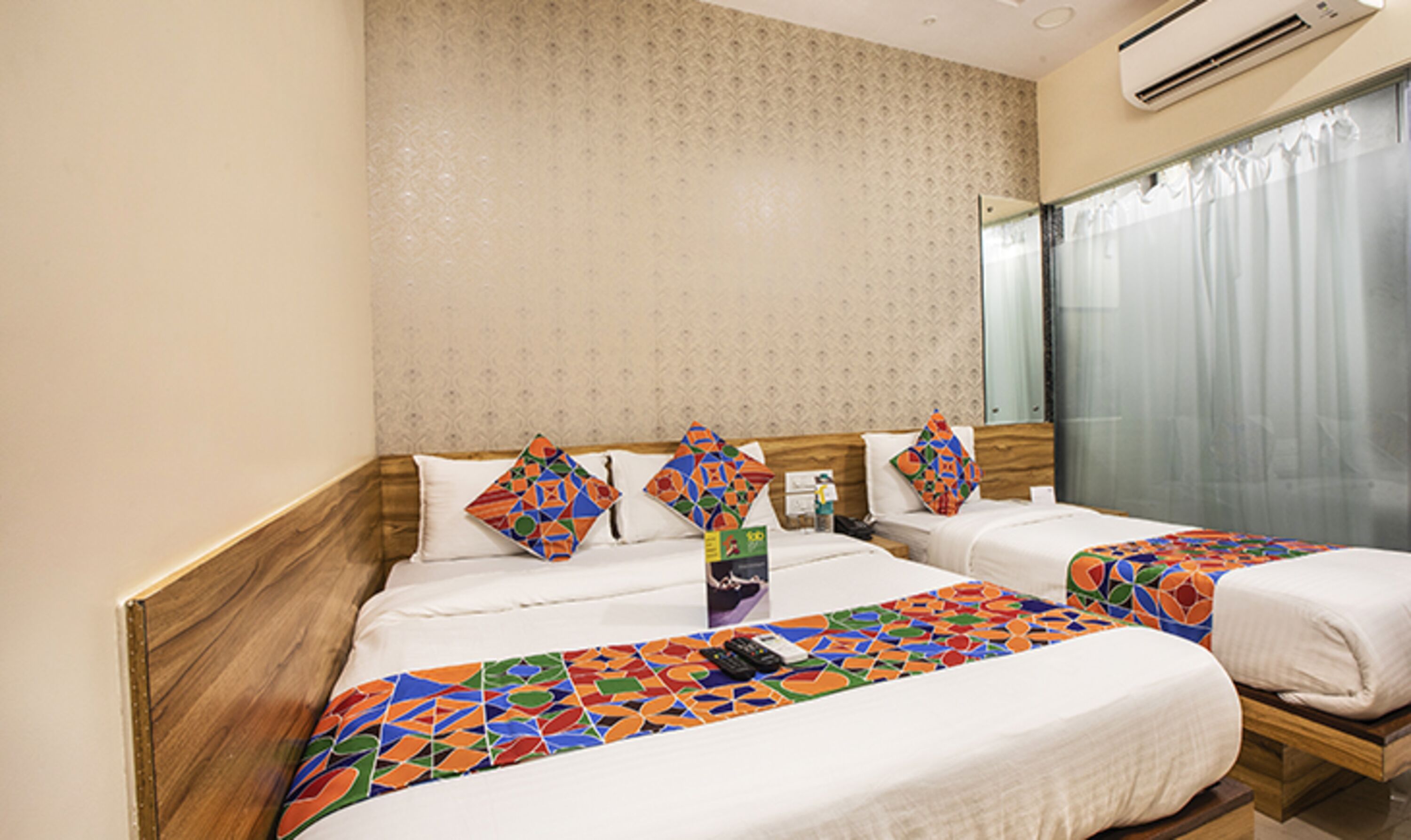 A hotel room with two beds, a wooden headboard, patterned wallpaper, and a large window with curtains.