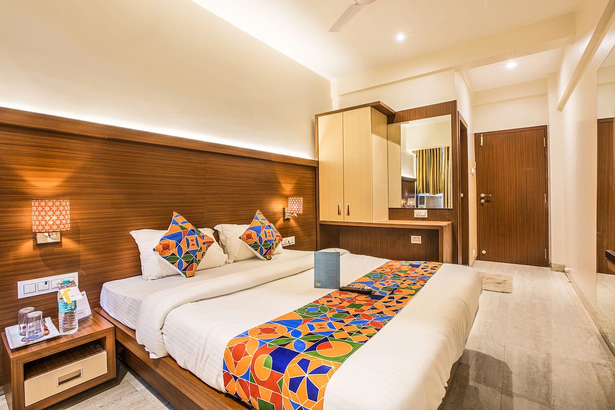 A modern hotel room with a large bed, wooden headboard, and a mirror above the bed. There is a nightstand with a lamp and a small table with a bottle and a glass.