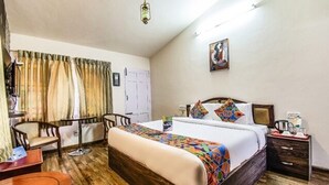 A hotel room with a wooden bed, a desk, a chair, a bedside table, and a painting on the wall. - Fabescape St. Clouds (Ootacamund)