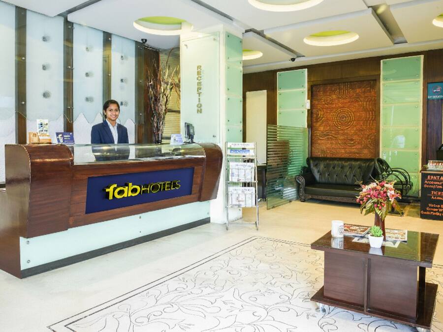 Een receptiebalie met een bord waarop 'fabHOTELS' staat en een receptionist. Er is een groene wand met een decoratief paneel en een zithoek met een groene bank.