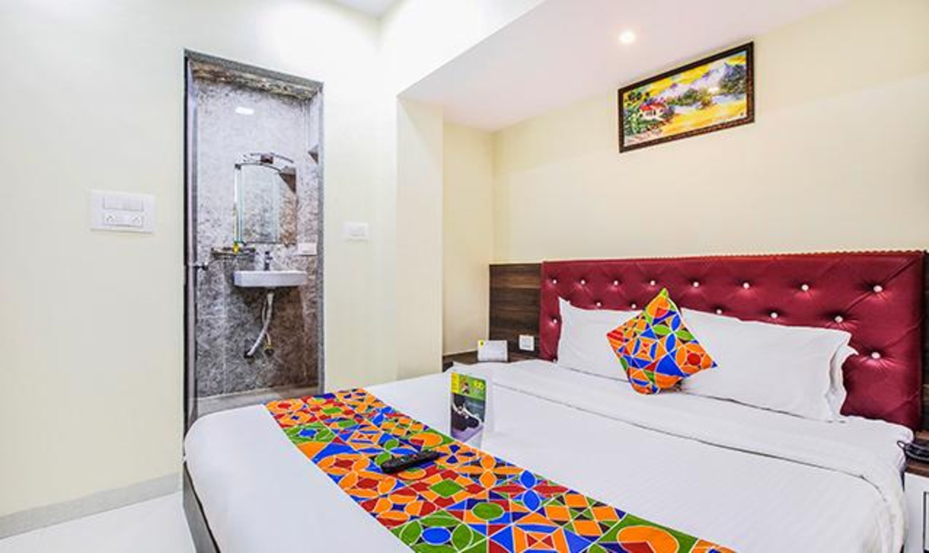 A hotel room with a bed, a red headboard, a colorful geometric bedspread, a white pillow, a nightstand, a framed picture on the wall, and a bathroom with a shower and sink.