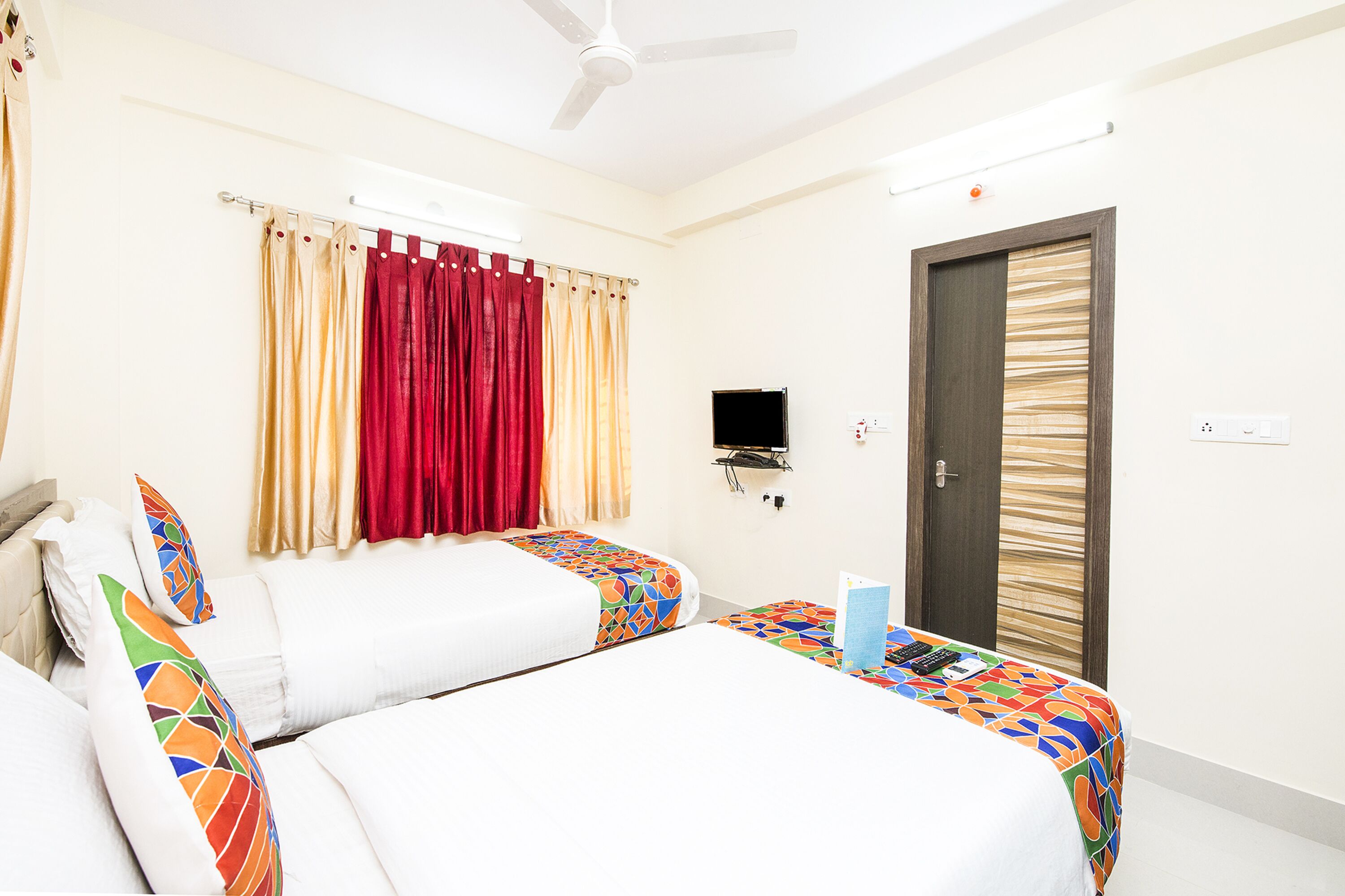 A hotel room with two beds, a ceiling fan, a TV, and a door with a patterned curtain.