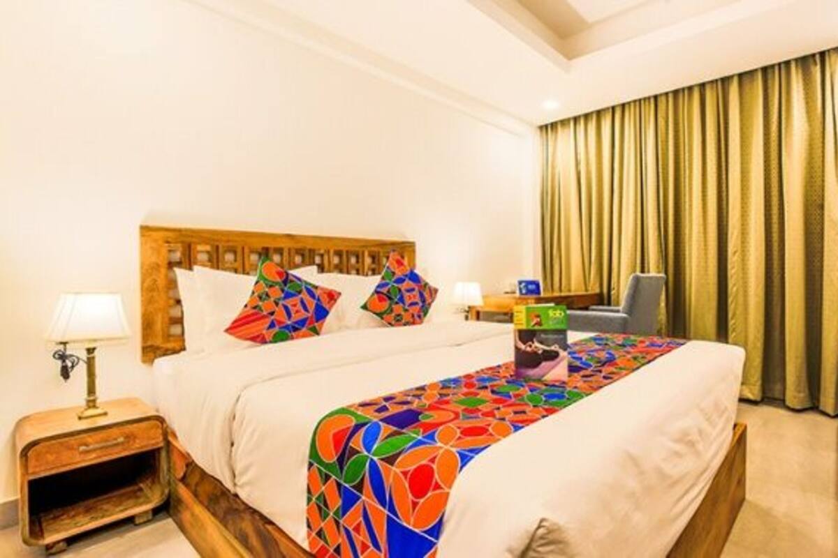 A hotel room with a large bed, a wooden headboard, a nightstand with a lamp, a chair, and a window with curtains.