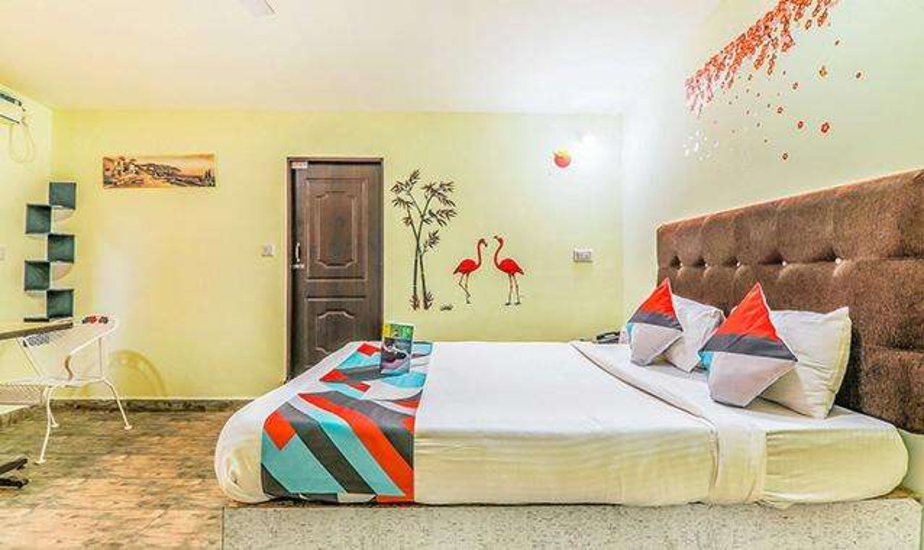 A bedroom with a bed, a door, a wall with a painting of flamingos, and a chair.