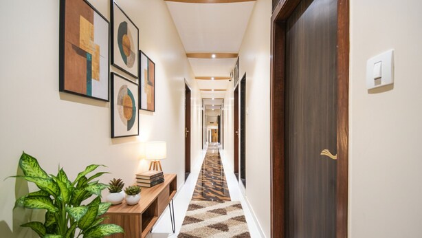 A hallway with a patterned carpet, framed artwork on the walls, a wooden console table with a lamp, and a potted plant. - FabHotel Orange Suites (Mumbai)